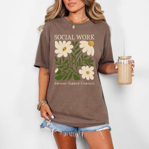 May include: Brown t-shirt with a floral design and the text "Social Work Advocate Support Empower".