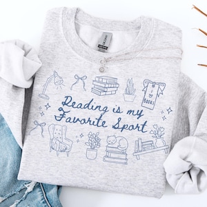Peut inclure: Un sweat-shirt gris clair avec l'inscription "Reading is my Favorite Sport" en bleu marine. Le motif comprend des illustrations de livres, une lampe, un chat et d'autres éléments liés à la lecture. Un collier argenté avec un pendentif en forme de fleur est drapé sur le sweat-shirt.