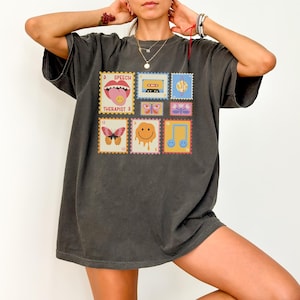 May include: A dark gray oversized t-shirt with a graphic print of colorful stamps featuring a smiley face, butterfly, music note, and the text "Speech Therapist".