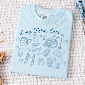 May include: Light blue t-shirt with the text "Long Term Care" and illustrations of medical supplies, including a wheelchair, walker, bandages, and a thermometer.