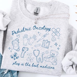 May include: Gray sweatshirt with a blue graphic design that says "Pediatric Oncology" and includes various medical symbols, such as a stethoscope, thermometer, and bandage. The text below the graphic says "play is the best medicine".