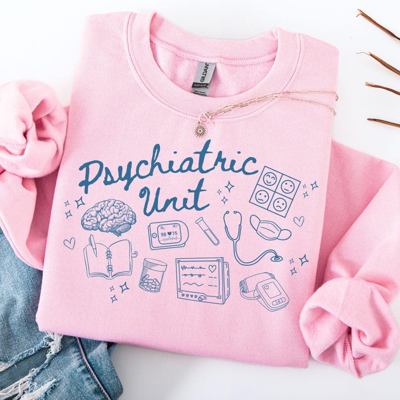 May include: Pink sweatshirt with the text "Psychiatric Unit" in blue lettering. The sweatshirt features a variety of blue illustrations including a brain, a stethoscope, a notebook, a pill bottle, a heart rate monitor, and a blood pressure cuff.