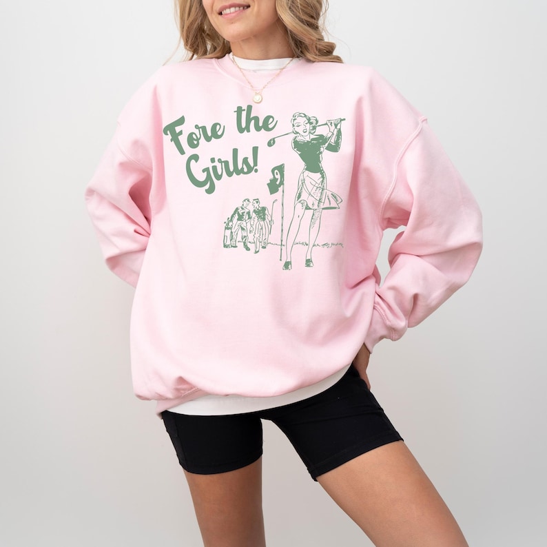 K&ouml;nnte beinhalten: Rosafarbener Pullover mit einer gr&uuml;nen Grafik einer Frau, die Golf spielt, und dem Text "Fore the Girls!"