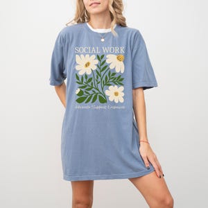 May include: A light blue t-shirt with a floral design and the text "Social Work Advocate Support Empower".