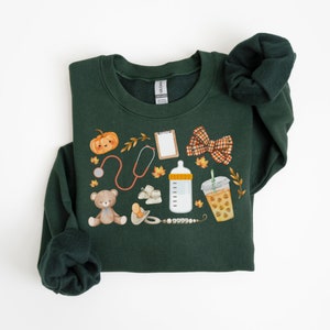 May include: A dark green sweatshirt with a cute autumn-themed design featuring a teddy bear, a pumpkin, a baby bottle, a stethoscope, a pacifier, a bow, a coffee cup, and a clipboard. The design is perfect for a new parent or someone who loves fall.
