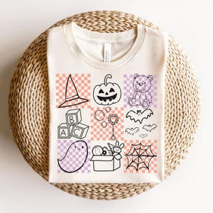 May include: Cream-colored t-shirt featuring a grid of Halloween-themed illustrations. The design includes a witch's hat, jack-o'-lantern, teddy bear, blocks, bubbles, bats, ghost, toys, and a spiderweb. The illustrations are outlined in black on a checkered background.