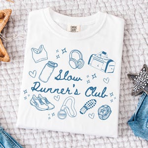 May include: White t-shirt with a blue graphic design that says "Slow Runner's Club". The graphic includes a water bottle, headphones, a sports bra, a gym bag, running shoes, a finishers medal, a watch, and hearts.