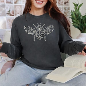 May include: Charcoal grey sweatshirt with a decorative bee motif, incorporating floral and leaf elements. The bee design includes the text "Bee like Jesus" and "Ephesians 5:1". This long-sleeved top is a relaxed, casual garment.