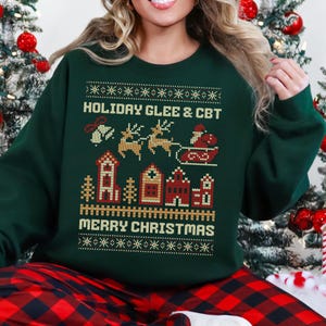 May include: A dark green Christmas sweatshirt with a festive design. The design includes the text "HOLIDAY GLEE & CBT" and "MERRY CHRISTMAS", along with images of reindeer, Santa's sleigh, houses, and trees. The sweatshirt is a cozy holiday garment.