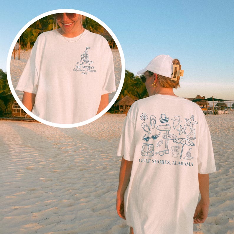 May include: White t-shirt with a blue graphic on the back featuring beach-themed illustrations and the text "GULF SHORES, ALABAMA." The front of the shirt has a smaller graphic and text. The shirt is worn on a beach.