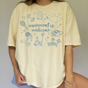 May include: A yellow t-shirt with a blue graphic that says "movement is medicine". The graphic includes illustrations of various fitness equipment, including a kettlebell, dumbbells, a yoga mat, a tennis racket, jump rope, a stationary bike, and a weight bench.