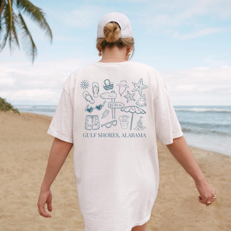 May include: White t-shirt with a blue graphic of beach-themed icons, including sunglasses, a starfish, and a beach umbrella. The text "GULF SHORES, ALABAMA" is printed below the graphic. The shirt is worn by a person on a sandy beach.