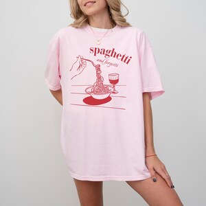May include: Pink oversized t-shirt with a red graphic of a hand holding a fork with spaghetti and a glass of wine. The text on the shirt reads "spaghetti and forgetti."