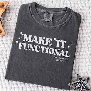 May include: Charcoal grey t-shirt featuring the white text "MAKE IT FUNCTIONAL" and "OCCUPATIONAL THERAPY." The shirt is folded on a white, textured surface. A label reads "COMFORT COLORS." The shirt is a medium size.