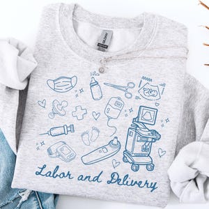May include: A pale grey sweatshirt featuring blue line drawings of medical equipment and baby-related symbols. The words "Labor and Delivery" are written in a script font. A silver necklace with a sunflower pendant is draped across the sweatshirt.