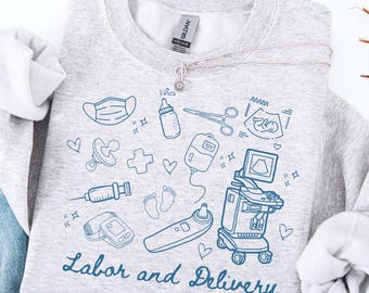 Può includere: Una felpa grigio chiaro con illustrazioni blu a tratto di strumenti medici e articoli per bambini. Le parole "Labor and Delivery" sono scritte in un carattere script. Una collana d'argento con un ciondolo a forma di girasole è drappeggiata sulla felpa.