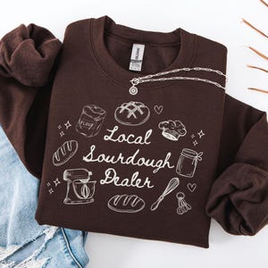 May include: Brown sweatshirt with a white graphic design that says "Local Sourdough Dealer". The graphic includes illustrations of bread, flour, a chef's hat, a mixer, and a whisk.