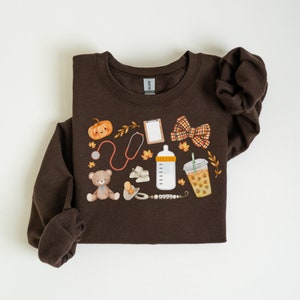 May include: A brown sweatshirt with a design featuring a teddy bear, a bottle, a stethoscope, a pumpkin, a coffee cup, a bow, and a pacifier. The design is in a fall theme with orange, brown, and yellow colors.