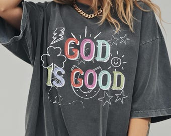 May include: Dark gray t-shirt with the phrase "GOD IS GOOD" in colorful, outlined letters. The design includes a lightning bolt, shamrock, stars, a horseshoe, and a smiling sun. The shirt has short sleeves and a relaxed fit.