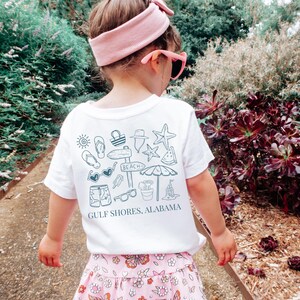 May include: A white t-shirt with a beach-themed graphic print in blue, featuring a sun, sunglasses, and a sign that says "Beach." The text "Gulf Shores, Alabama" is printed below the graphic. The child is wearing a pink headband and sunglasses.