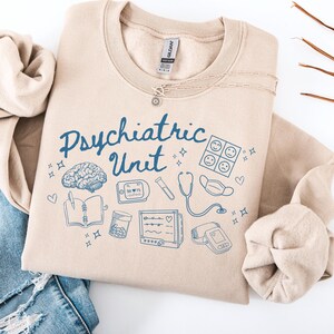 May include: Tan sweatshirt with the text "Psychiatric Unit" in blue lettering. The sweatshirt features a variety of blue illustrations including a brain, a stethoscope, a heart monitor, and pills.