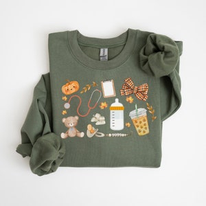 May include: A green sweatshirt with a fall-themed design featuring a pumpkin, stethoscope, baby bottle, teddy bear, pacifier, and a cup of iced coffee. The design is in watercolor style.