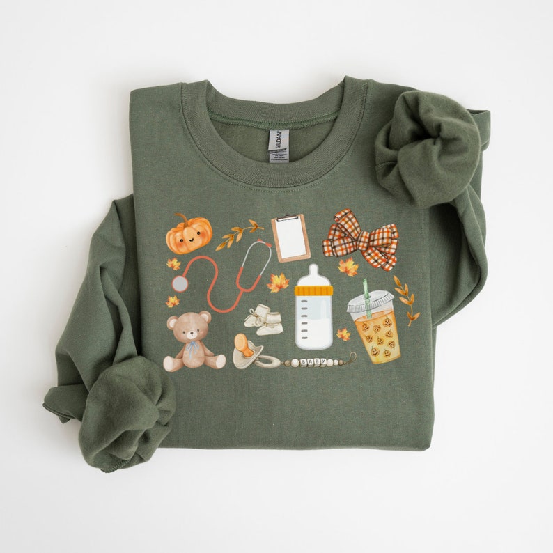 May include: A green sweatshirt with a fall-themed design featuring a pumpkin, stethoscope, baby bottle, teddy bear, pacifier, and a cup of iced coffee. The design is in watercolor style.