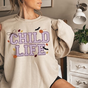 May include: A beige crewneck sweatshirt featuring the text "CHILD LIFE" in large, purple and white gingham-patterned block letters. The design is decorated with small orange pumpkins, black bats, and scattered autumn leaves in red, yellow, and brown.