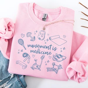 Pode incluir: Camisola cor-de-rosa com o texto "movement is medicine" em azul. A camisola apresenta uma variedade de &iacute;cones de fitness, incluindo um haltere, um kettlebell, um tapete de ioga, uma corda de saltar, uma bicicleta de exerc&iacute;cio e um banco de muscula&ccedil;&atilde;o.