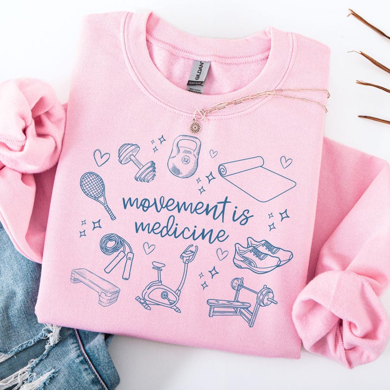 Pode incluir: Camisola cor-de-rosa com o texto "movement is medicine" em azul. A camisola apresenta uma variedade de &iacute;cones de fitness, incluindo um haltere, um kettlebell, um tapete de ioga, uma corda de saltar, uma bicicleta de exerc&iacute;cio e um banco de muscula&ccedil;&atilde;o.