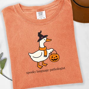 May include: Orange Comfort Colors t-shirt featuring a cartoon goose wearing a witch hat and scarf, holding a jack-o'-lantern. The text "spooky language pathologist" is printed below the goose. The shirt has a crew neck and a tag.