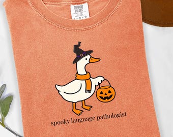 Può includere: T-shirt arancione Comfort Colors con un'oca dei cartoni animati con cappello da strega e sciarpa, con in mano una lanterna a forma di zucca. Il testo "spooky language pathologist" è stampato sotto. Girocollo ed etichetta.