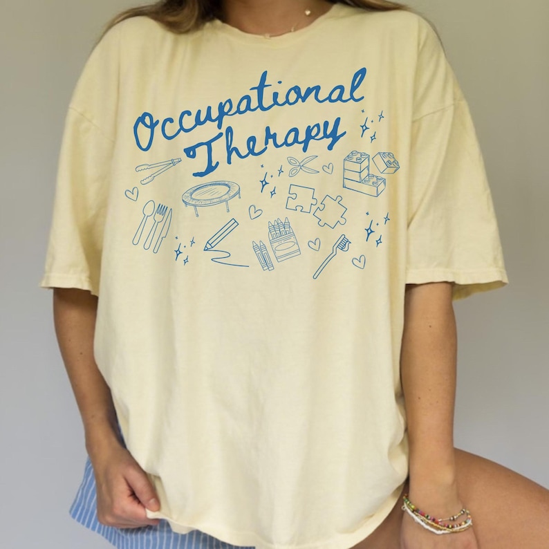 May include: A light yellow t-shirt with the text "Occupational Therapy" in blue script. The shirt features a variety of blue illustrations of objects related to occupational therapy, including a pencil, scissors, a toothbrush, a puzzle, a trampoline, and a box of crayons.