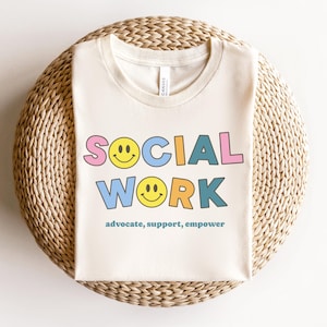 May include: A beige t-shirt with the text "SOCIAL WORK" in colorful letters with smiley faces. The text "advocate, support, empower" is printed below the main text.