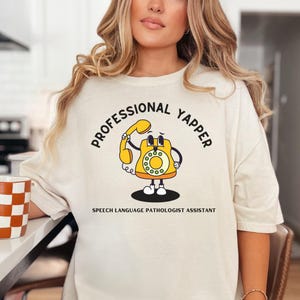 May include: A white t-shirt with the text "Professional Yapper" in a black circle around a cartoon yellow rotary phone. The text "Speech Language Pathologist Assistant" is printed below the phone.
