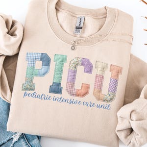 May include: A beige crewneck sweatshirt featuring the letters PICU in a patchwork design with various pastel-colored fabrics, including gingham, floral, and solid patterns. Below the letters, the text pediatric intensive care unit is printed in blue.