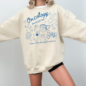 May include: A beige sweatshirt with a blue graphic design that says "Oncology" and includes medical symbols such as a stethoscope, syringe, thermometer, and pills. The text below the graphic says "Hope is the strongest medicine."