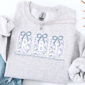 May include: A light gray sweatshirt with the letters "RBT" in white, framed by floral designs and light blue bows. The words "Registered Behavior Technician" are written below the letters in a script font. A silver necklace with a flower pendant is draped across the sweatshirt.