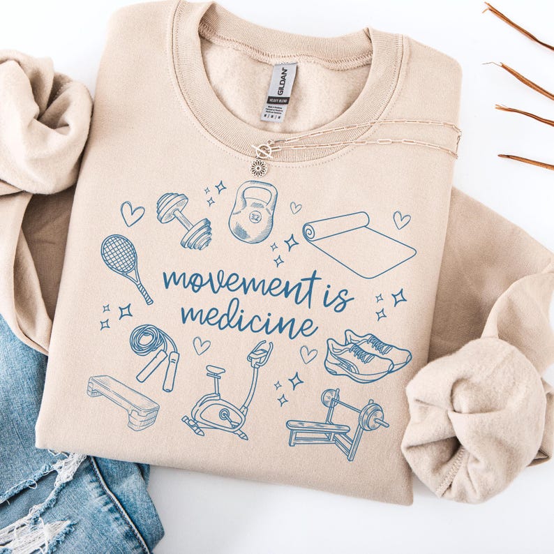 Pode incluir: Uma camisola bege com o texto "movement is medicine" em letras azuis. A camisola apresenta uma variedade de &iacute;cones de fitness em azul, incluindo um haltere, um tapete de ioga, um kettlebell, uma corda de saltar, uma bicicleta de exerc&iacute;cio e um par de sapatos de corrida.