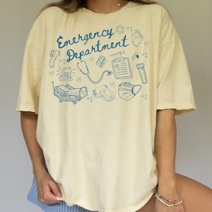 May include: A light yellow t-shirt with a blue graphic design that says "Emergency Department" and features medical symbols such as a stethoscope, a heart rate monitor, a syringe, and a face mask.