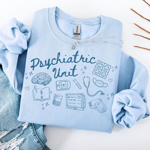 May include: Light blue sweatshirt with the text "Psychiatric Unit" in a blue script font. The sweatshirt features a variety of blue illustrations related to mental health, including a brain, a stethoscope, a pill bottle, a notebook, and a heart rate monitor.