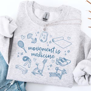 Pode incluir: Camisola de moletom cinza com um design gr&aacute;fico azul de v&aacute;rios equipamentos de gin&aacute;stica, incluindo um kettlebell, halteres, um tapete de ioga, uma corda de pular, uma bicicleta ergom&eacute;trica, uma barra de peso, uma plataforma de step, t&ecirc;nis de corrida e uma raquete de t&ecirc;nis. O texto "movement is medicine" est&aacute; escrito em azul.