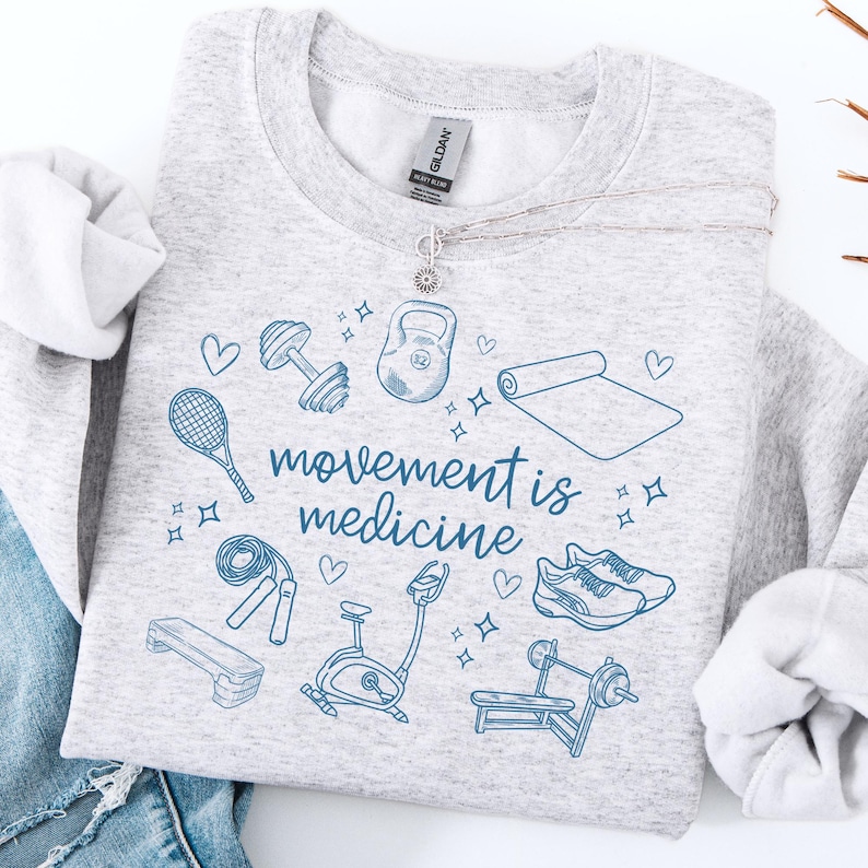 Pode incluir: Camisola de moletom cinza com um design gr&aacute;fico azul de v&aacute;rios equipamentos de gin&aacute;stica, incluindo um kettlebell, halteres, um tapete de ioga, uma corda de pular, uma bicicleta ergom&eacute;trica, uma barra de peso, uma plataforma de step, t&ecirc;nis de corrida e uma raquete de t&ecirc;nis. O texto "movement is medicine" est&aacute; escrito em azul.
