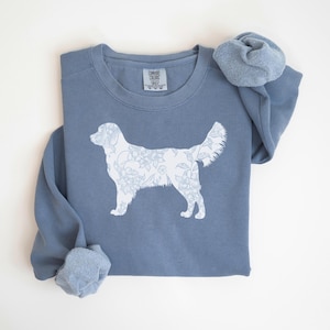 Op de afbeelding: Een stoffig blauw sweatshirt met een witte bloemenhond-silhouet. Het sweatshirt heeft een ronde hals en lange mouwen. Het Comfort Colors-label is zichtbaar bij de halslijn. Het hondenontwerp is gevuld met een bloemenpatroon.
