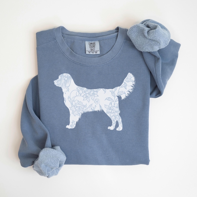 Op de afbeelding: Een stoffig blauw sweatshirt met een witte bloemenhond-silhouet. Het sweatshirt heeft een ronde hals en lange mouwen. Het Comfort Colors-label is zichtbaar bij de halslijn. Het hondenontwerp is gevuld met een bloemenpatroon.