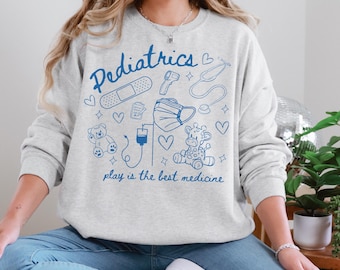 May include: A gray sweatshirt with a blue graphic design that says "Pediatrics" and includes medical symbols like a stethoscope, thermometer, bandage, and IV bag. The graphic also includes a teddy bear, a giraffe, and the text "play is the best medicine."