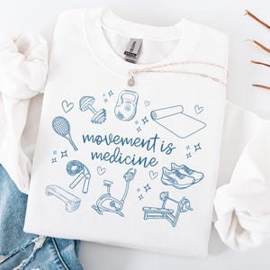 Pode incluir: Camisola branca com uma estampa gr&aacute;fica azul de v&aacute;rios equipamentos de gin&aacute;stica e o texto "movement is medicine".