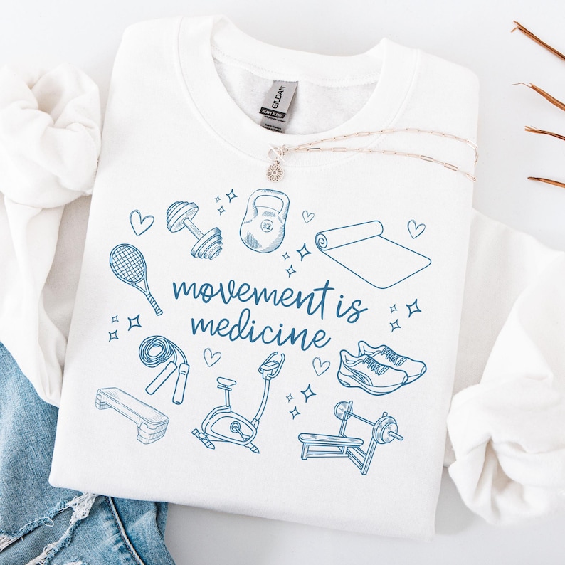 Pode incluir: Camisola branca com uma estampa gr&aacute;fica azul de v&aacute;rios equipamentos de gin&aacute;stica e o texto "movement is medicine".