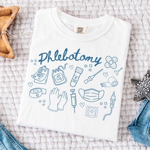 May include: White t-shirt with a blue doodle design that says "Phlebotomy" and includes medical symbols like a blood bag, syringe, bandage, and face mask.