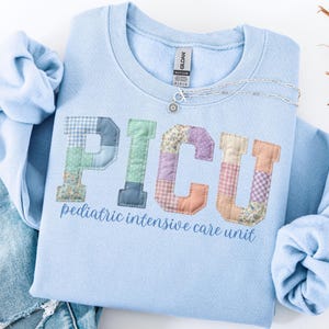 May include: A light blue crewneck sweatshirt featuring the letters PICU in a patchwork design with various floral and gingham patterns. Below the letters, the text pediatric intensive care unit is printed in a matching blue script font.
